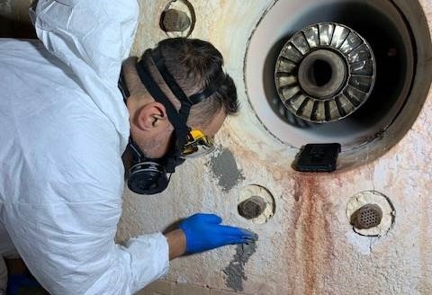 A person wearing a safety suit and ventilator inspects a piece of equipment up close.