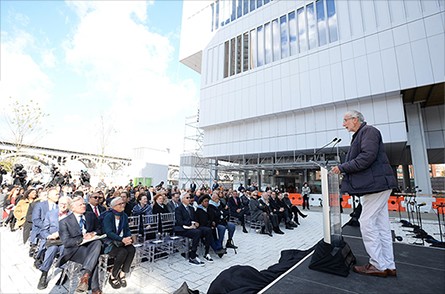 A person at a podium addreses a seated audience outside at The Plaza with Lenfest Center for the Arts adjacent to the audience.