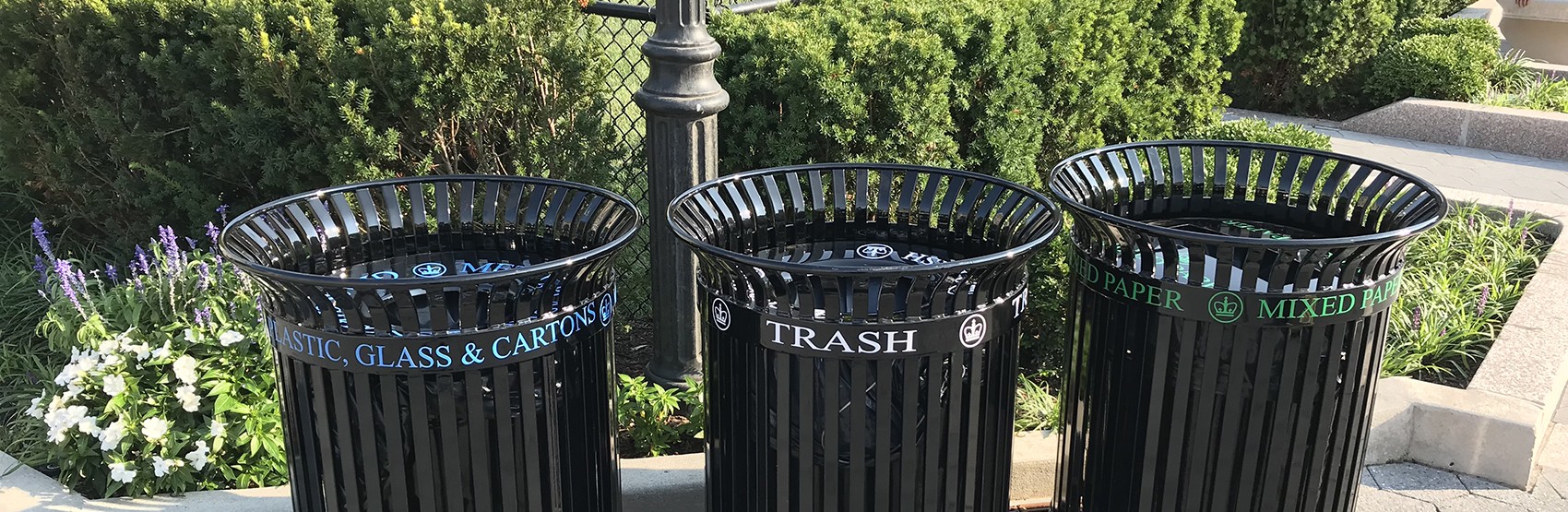 Three black bins for mixed paper, trash, and glass, metal, and plastic sit on the corner of a pathway