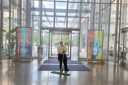 A Facilities and Operations custodial member stands in the Jerome L. Greene Science Center lobby, with two framed pieces of art behind her.