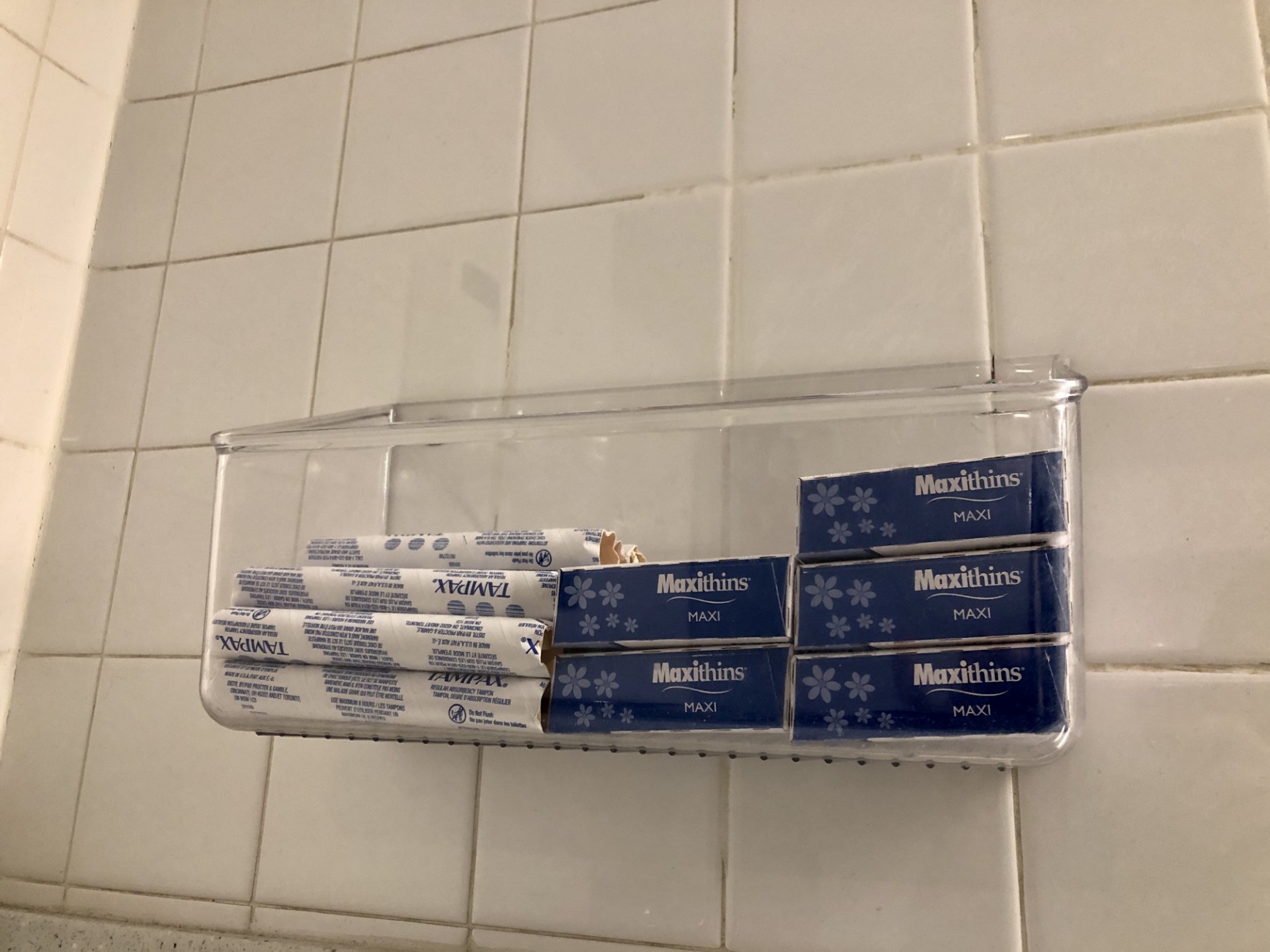 A plastic container on the wall of a restroom that has free tampons and menstraul pads.