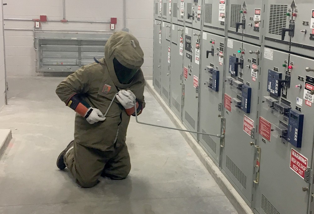 Someone in a electrical safety suit and helmet makes adjustments to equipment using a very long crank.