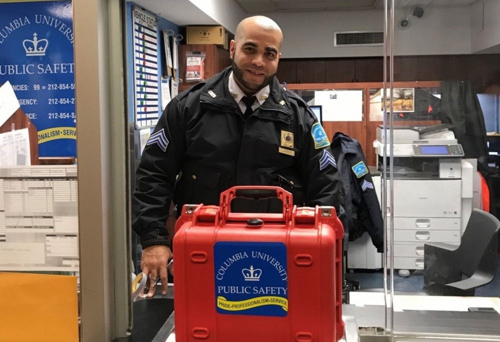 A Public Safety officer stands at the welcome desk at the Morningside Operations center with a red box containing an automated external defibrillator.