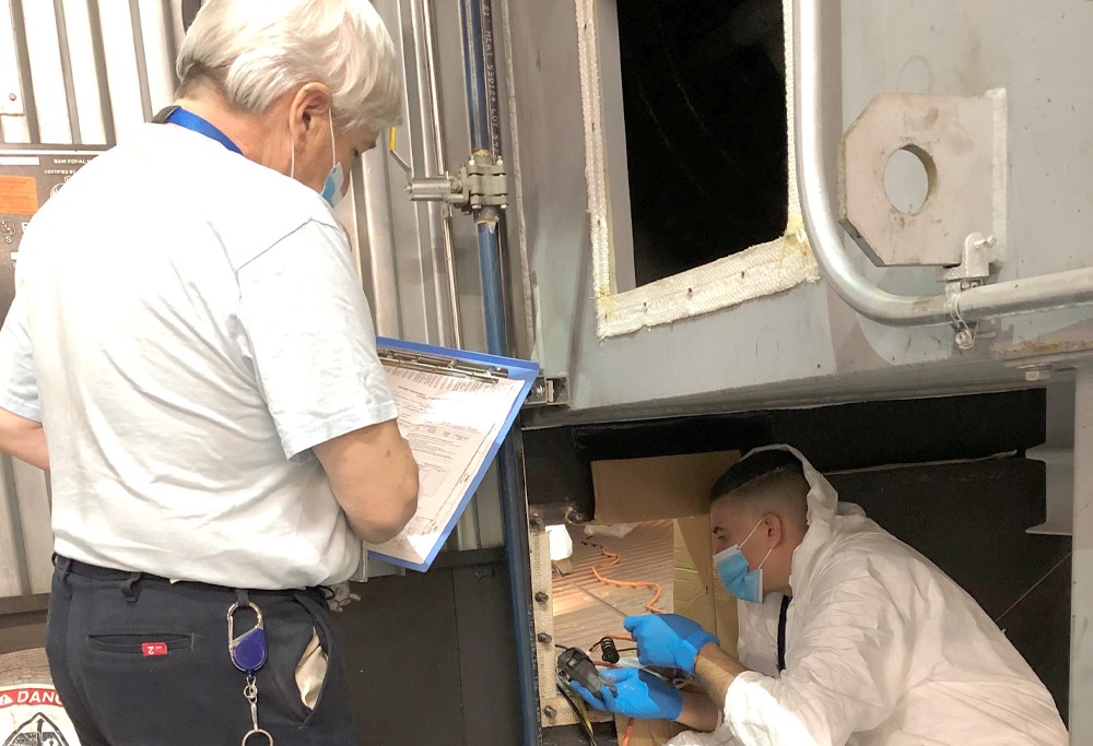 A person with a checklist monitors someone in a safety suit performing work in a duct.