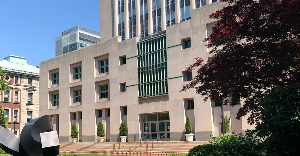 An exterior shot of Uris Hall on a cloudless day, with the curl sculpture on the lawn and a Japanese maple to the right of the building