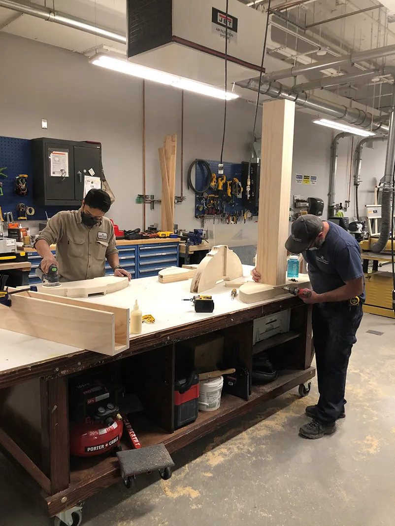 Two carpenters at a work bench assembling components of the custom frame for the artwork.  