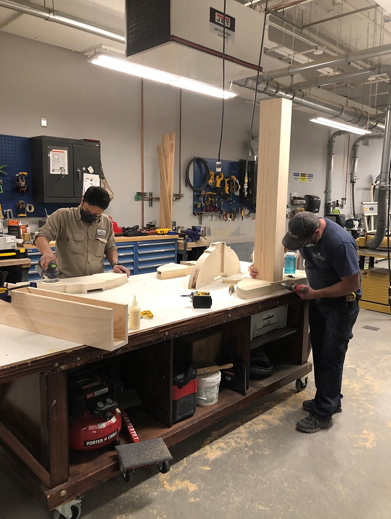 Two carpenters at a work bench assembling components of the custom frame for the artwork.  