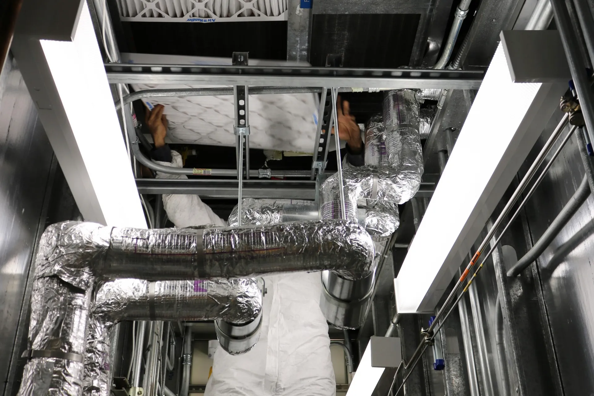 A Facilities and Operations employee wearing a white suit installs an air filter inside of a mechanical room.