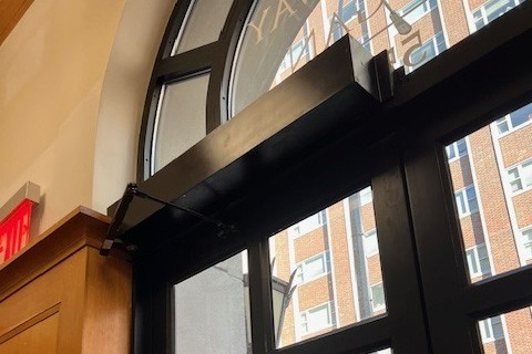 New accessible operators have been installed at the front entrance of Broadway Hall. A black accessible door operator that has been installed at the top of a door.